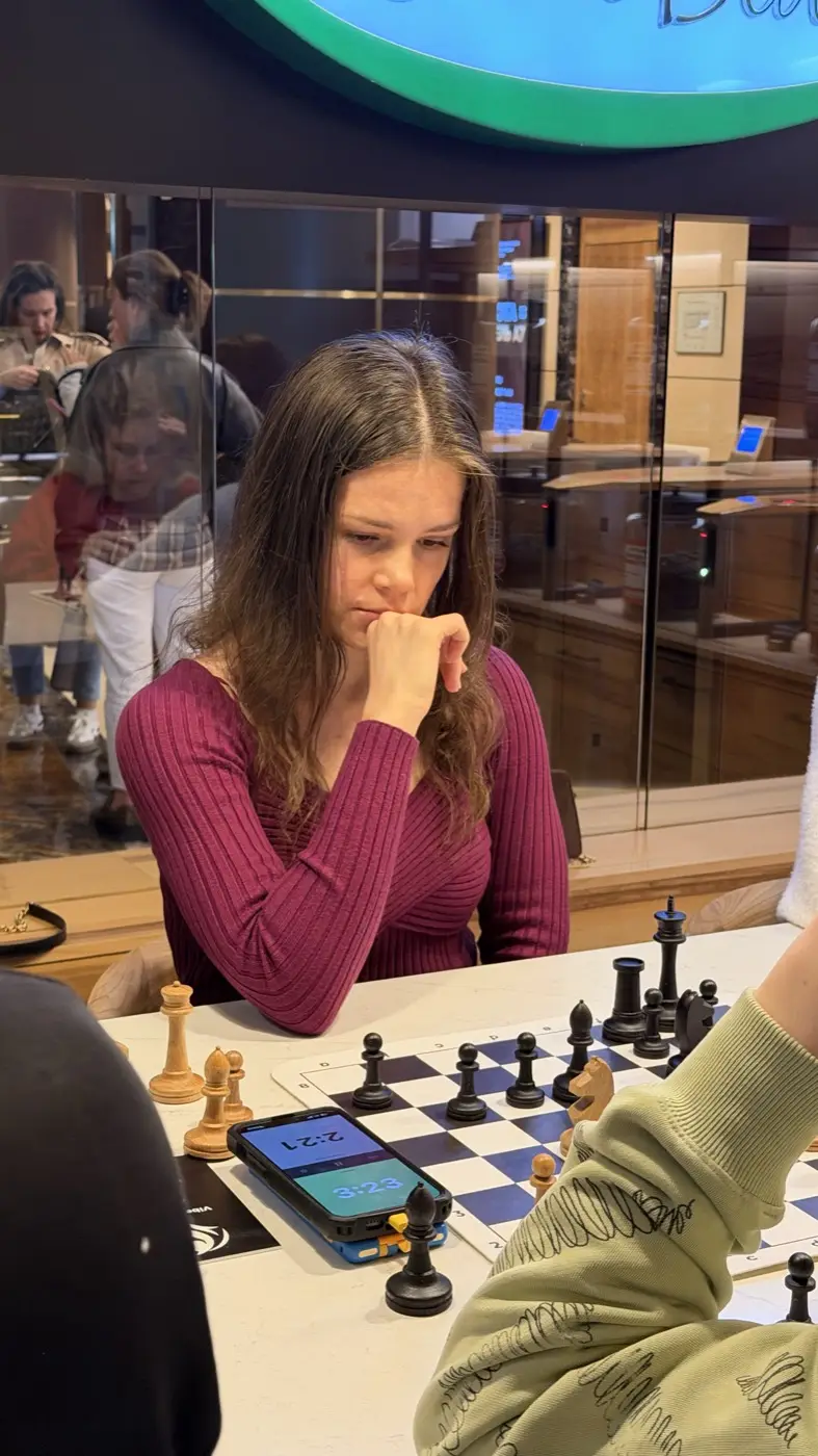 A player in a burgundy sweater thinking over a move, a phone clock beside her