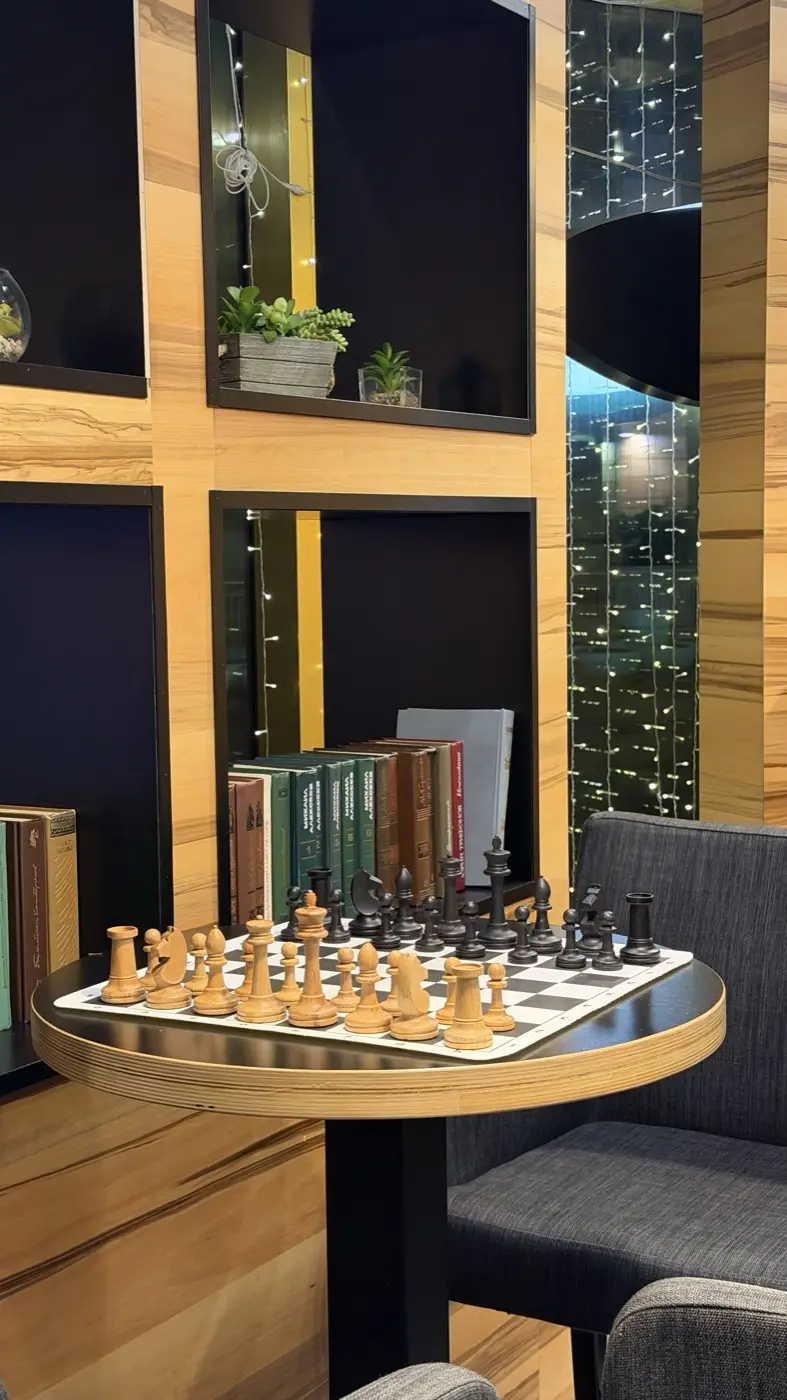 A chessboard on a small table next to a bookshelf and a window